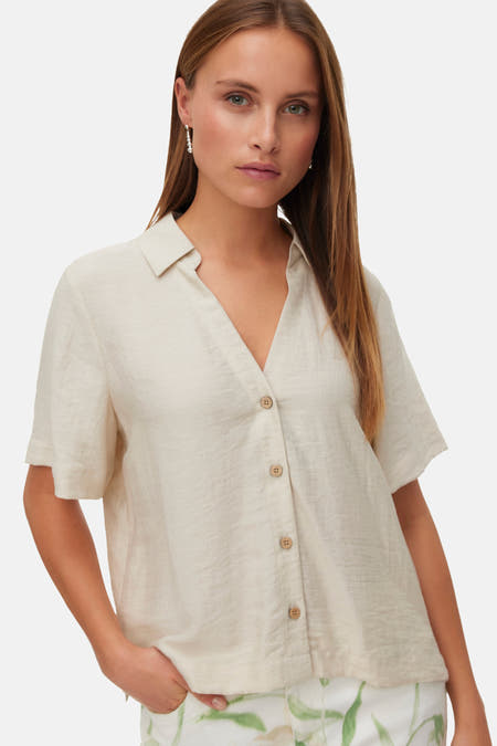 Chemise écru à manches courtes de Vero Moda, avec fermeture boutonnée et portée avec un pantalon blanc à imprimé floral.