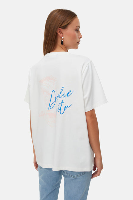 T-shirt blanc VERO MODA avec imprimé rose clair sur la poitrine, porté avec un jean bleu clair et des ballerines pointues bleu clair.