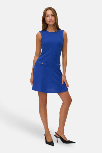 Model in blauwe tweed jurk met zwarte pumps op witte achtergrond.
