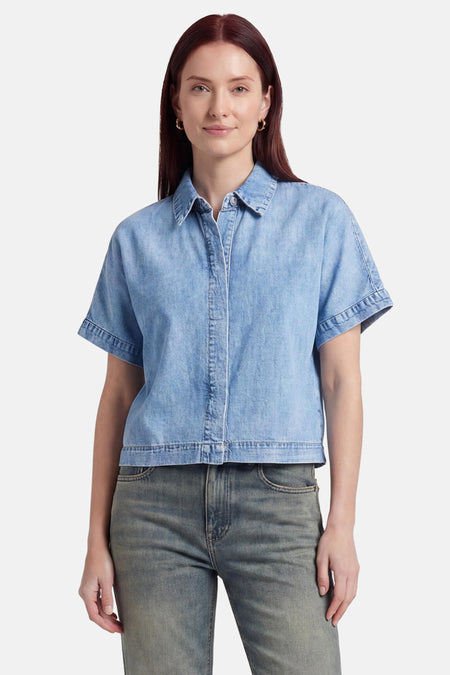 Chemisier en jean bleu clair à manches courtes de VERO MODA® avec col, fermeture boutonnée et coupe décontractée, porté avec un jean.