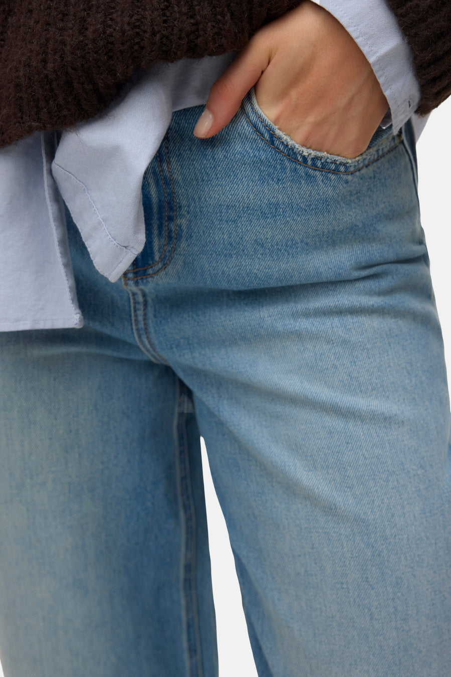 Jean bleu clair de VERO MODA, gros plan sur une main dans la poche à bord effiloché, porté avec un pull marron et une chemise bleue.