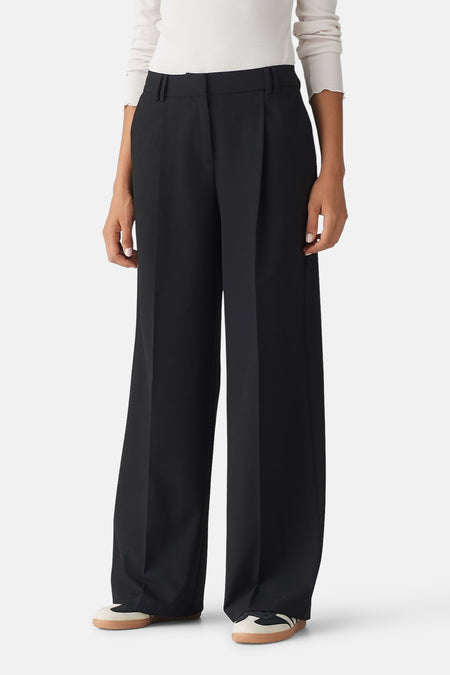 Pantalon noir OPUS avec taille élastique, poches latérales et jambes larges, dos, porté avec un haut blanc et des chaussures marron.