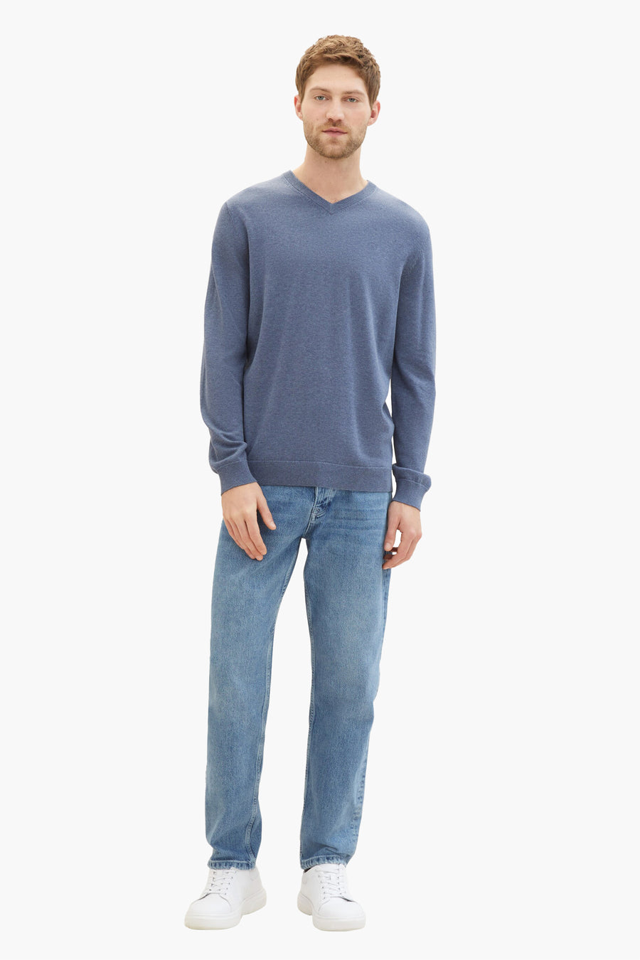 Man staat, blauwe trui, jeans, witte sneakers.