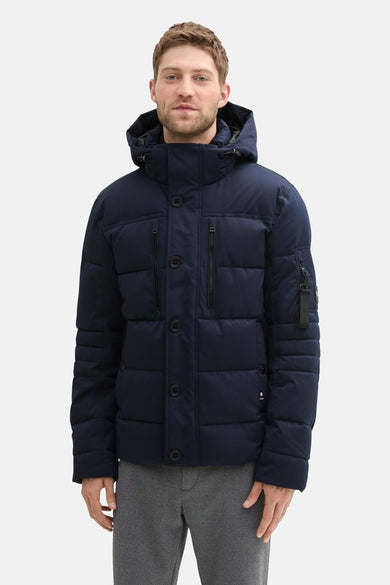 Manteau bleu court de TOM TAILOR, avec capuche, fermeture éclair et boutons, associé à un pantalon gris et des chaussures noires.