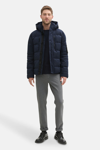 Korte blauwe jas van TOM TAILOR, met capuchon, rits- en knoopsluiting, gecombineerd met grijze broek en zwarte schoenen.