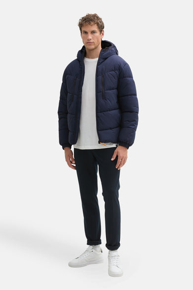 Korte blauwe jas van TOM TAILOR, met capuchon, gedragen over een wit T-shirt en donkere broek met witte sneakers.
