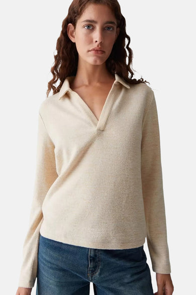 Vrouw met golvend bruin haar draagt een beige sweater met kraag en blauwe jeans.