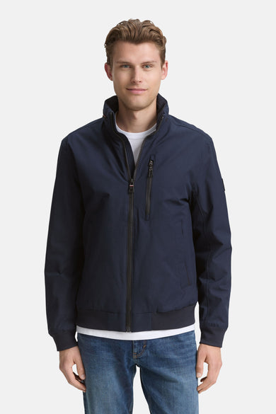 Manteau court bleu de TOM TAILOR, avec une fermeture éclair et un col montant, porté sur un t-shirt blanc et des jeans bleus.