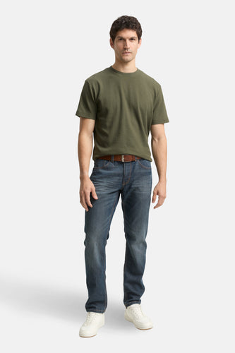 Man in olijfgroen T-shirt, jeans en witte sneakers staand tegen witte achtergrond.