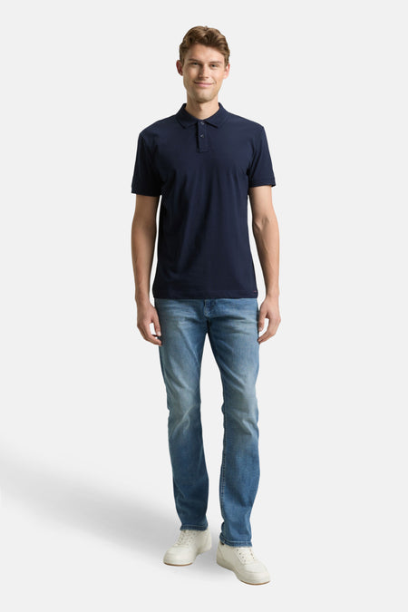 Man, staand, marineblauwe polo, blauwe jeans, witte sneakers.
