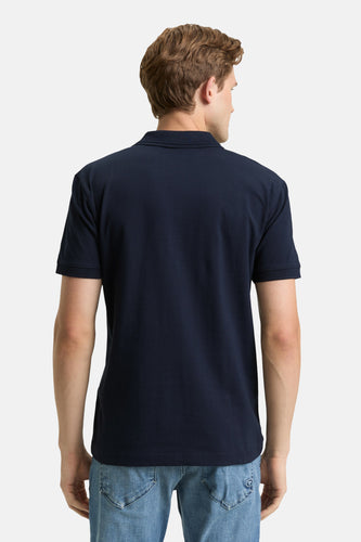 Man achterkant, marineblauw poloshirt, jeans.
