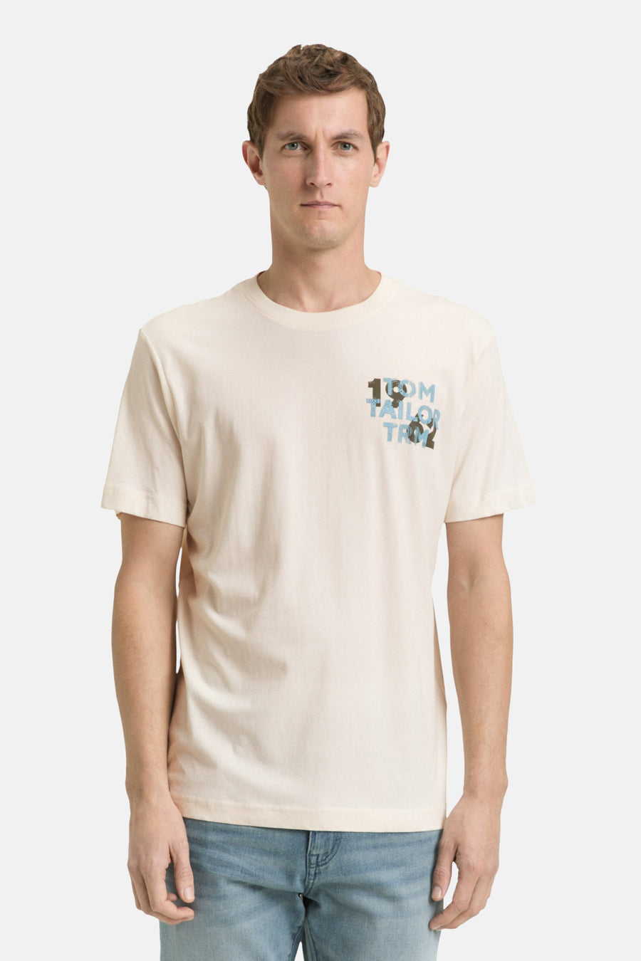 T-shirt écru à manches courtes de TOM TAILOR, avec une petite impression bleue en haut à gauche, associé à un jean et des baskets blanches.
