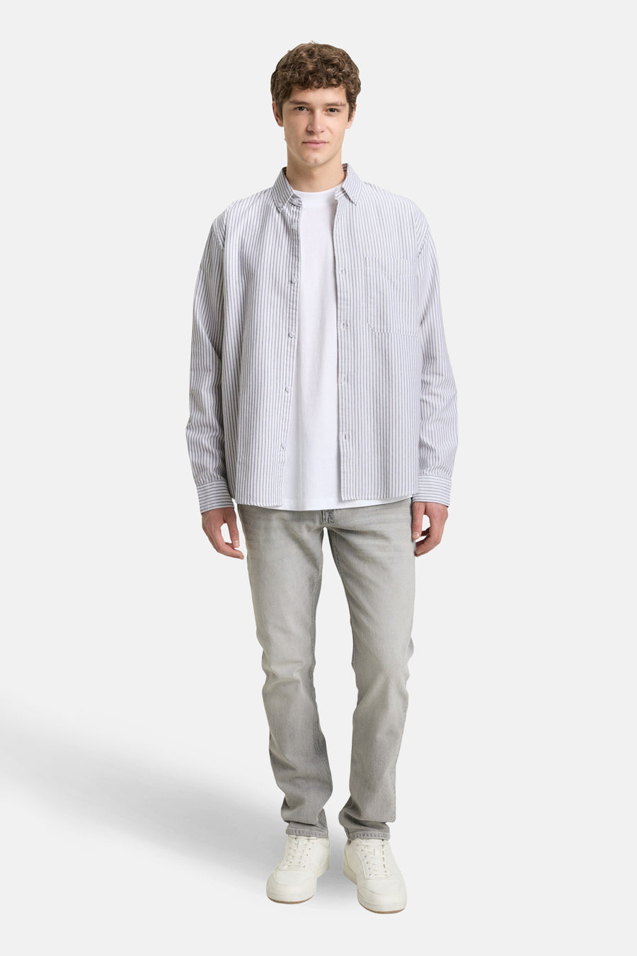 Staande man in wit t-shirt, gestreept overhemd, grijze jeans en witte sneakers.