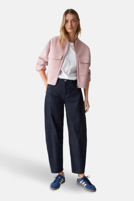 Een vrouw draagt een roze jas, een wit T-shirt, wijde donkerblauwe jeans en blauwe Adidas sneakers.