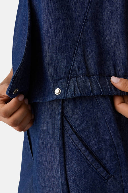 Chemisier en jean bleu foncé OPUS, sans manches, avec boutons-pression, taille élastique et poches latérales, tenu par des mains.