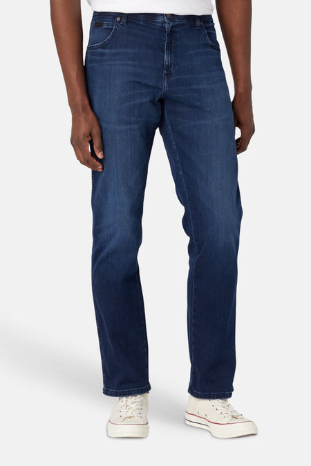 Vue arrière d'un jean droit bleu foncé Wrangler® avec écusson noir et coutures en « W » sur les poches arrière, porté avec des baskets blanches.