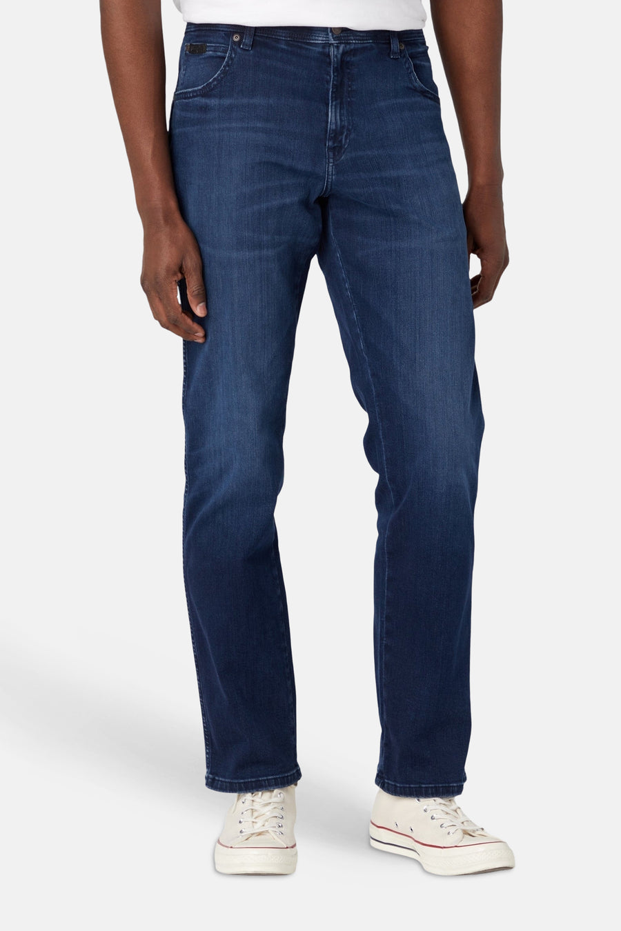 Vue arrière d'un jean droit bleu foncé Wrangler® avec écusson noir et coutures en « W » sur les poches arrière, porté avec des baskets blanches.