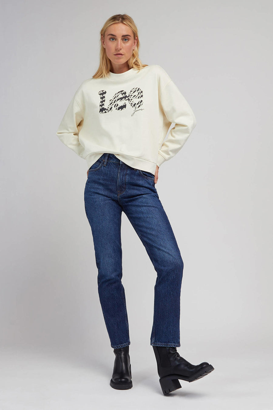 Vrouw in crèmekleurige trui met jeans en zwarte laarzen.
