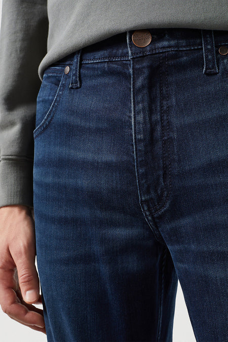 Jean droit bleu foncé de Wrangler, avec fermeture à boutons et à glissière, et un pull gris.