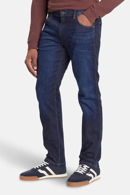 Jean droit en denim bleu marine de Wrangler, arrière-plan montré, associé à un t-shirt marron et des baskets.