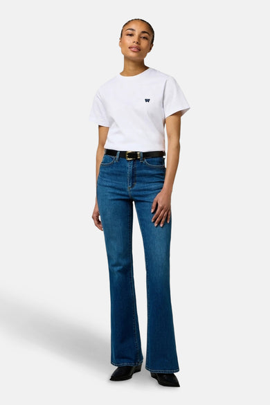 Jean Wrangler en denim bleu délavé, avec une ceinture noire et un T-shirt blanc à logo.