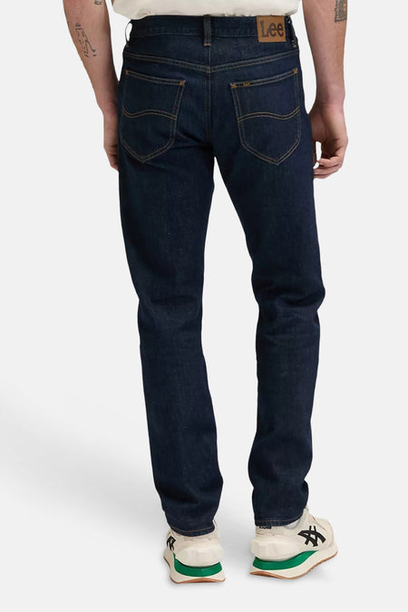 Donkere blauwe tapered jeans van Lee® met goudkleurige naden, achterzakken en rits, weergegeven van achteren op een model in wit shirt en witte sneakers met groene zool.