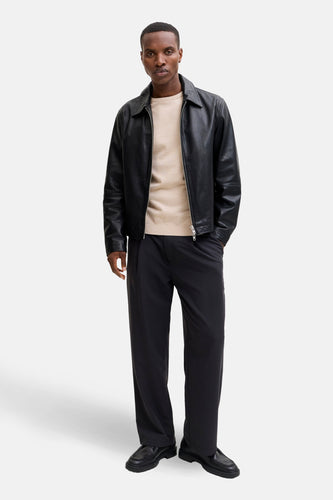 Een man die een zwarte leren jas, beige trui en zwarte broek draagt.