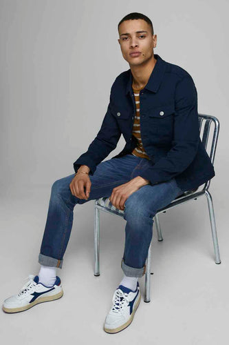 Zittende man in blauw jasje, gestreepte top, jeans en witte sneakers.