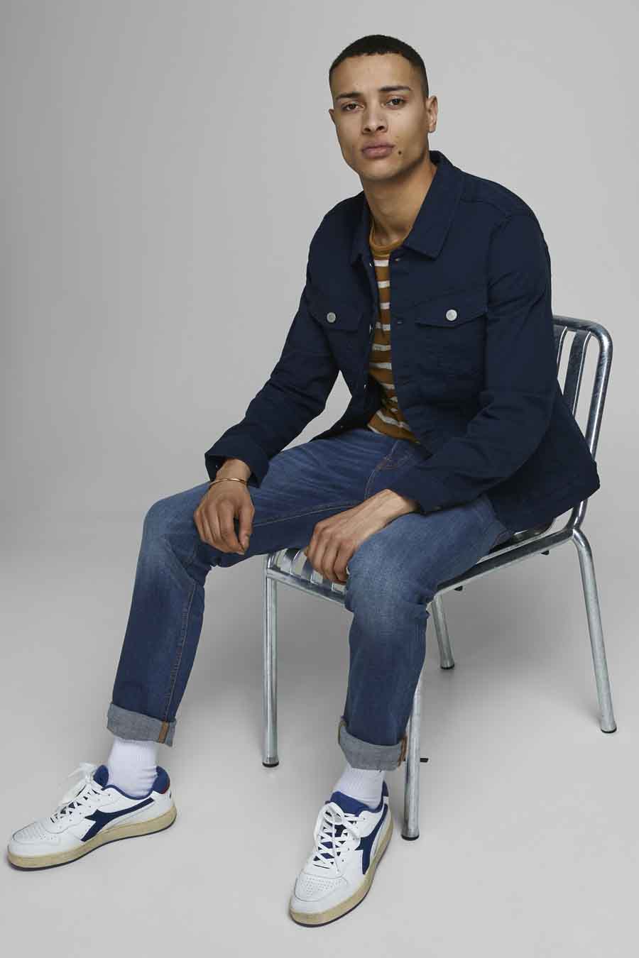 Zittende man in blauw jasje, gestreepte top, jeans en witte sneakers.