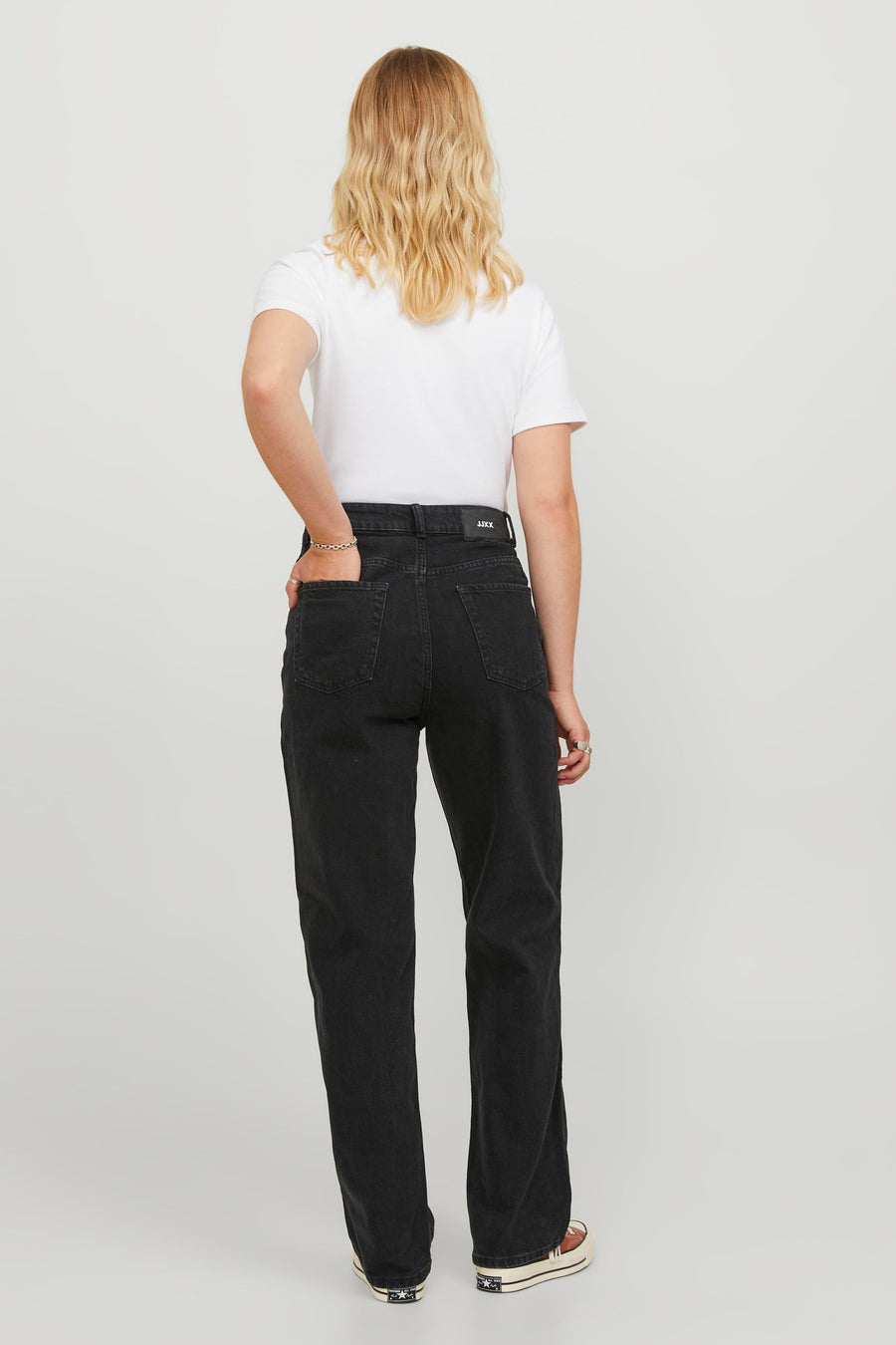 Model van achteren met zwarte spijkerbroek, wit T-shirt en Converse All Stars schoenen op een witte achtergrond.
