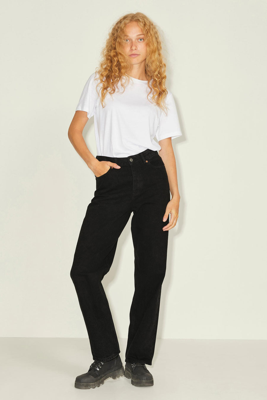 Model met blond krullend haar, witte t-shirt, zwarte jeans en zwarte boots, poseert voor een witte achtergrond.