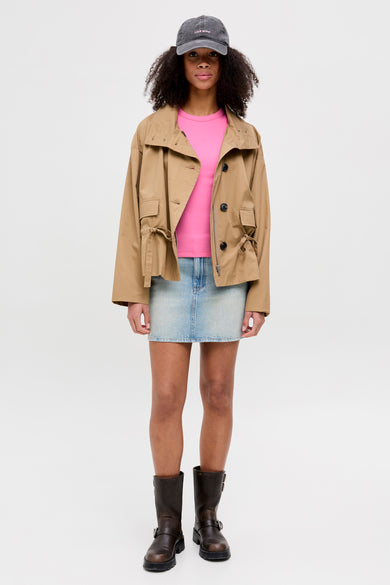 T-shirt rose de JJXX, porté sous un manteau brun et une jupe en jeans bleu, avec des bottes en cuir noires et un chapeau gris.