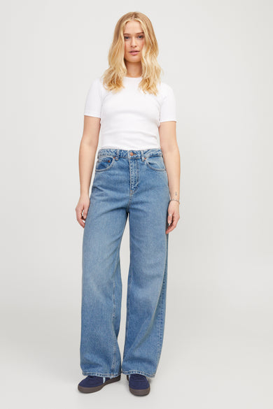 Jean décontracté en denim bleu moyen de JJXX, porté par un modèle en une chemise blanche et des chaussures bleues.