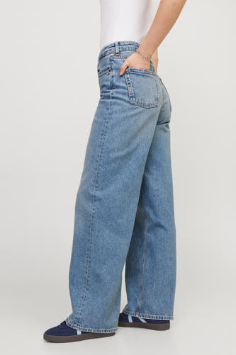 Jean en denim bleu moyen de JJXX, avec une main dans la poche et des chaussures à rayures bleues.