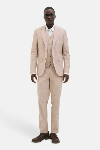 Man in beige driedelig pak met bruine schoenen, staand voor een witte achtergrond.