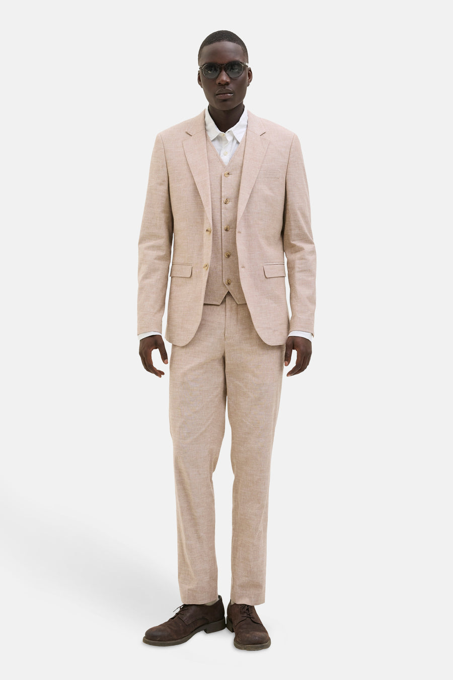 Man in beige driedelig pak met bruine schoenen, staand voor een witte achtergrond.