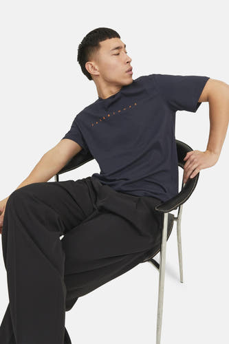 T-shirt bleu à manches courtes d'ORIGINALS par JACK & JONES, avec logo orange imprimé, porté par un modèle sur une chaise.