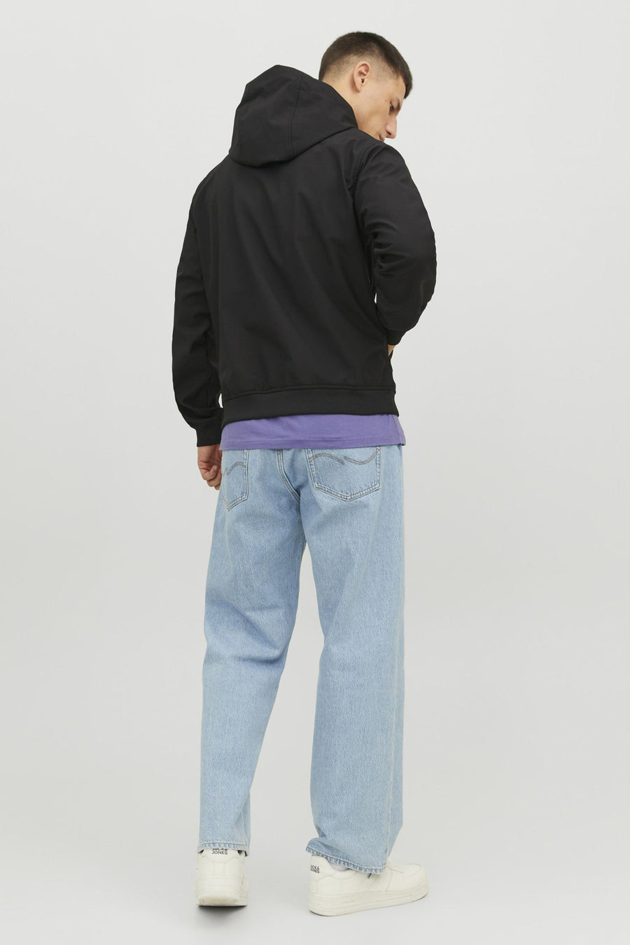 Man van achteren in een zwarte jas, paars t-shirt, wijde blauwe jeans en witte sneakers.
