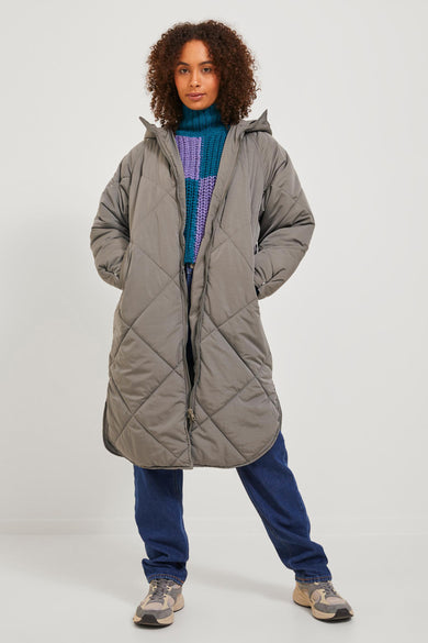 Manteau long marron de JJXX, avec un motif à carreaux, une capuche et porté avec un pull bleu et des jeans.
