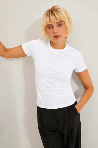 T-shirt blanc à manches courtes de JJXX, porté avec un pantalon noir, appuyé contre un mur blanc.