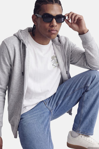 Sweat à capuche zippé gris de CORE BY JACK & JONES, avec capuche et poche avant, porté sur un t-shirt blanc avec imprimé dragon, jean bleu et baskets blanches, photo lifestyle.