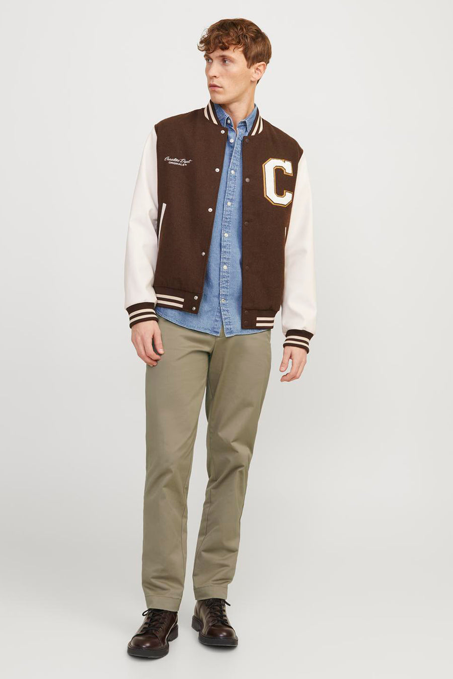 Beige chino van JACK & JONES JEANS INTELLIGENCE, gecombineerd met een denim overhemd, varsity jacket en bruine schoenen.