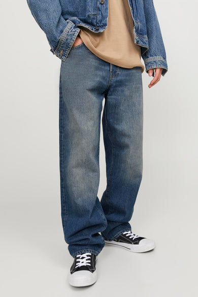 Persoon in denim jack, bruin shirt, blauwe jeans en zwarte sneakers.
