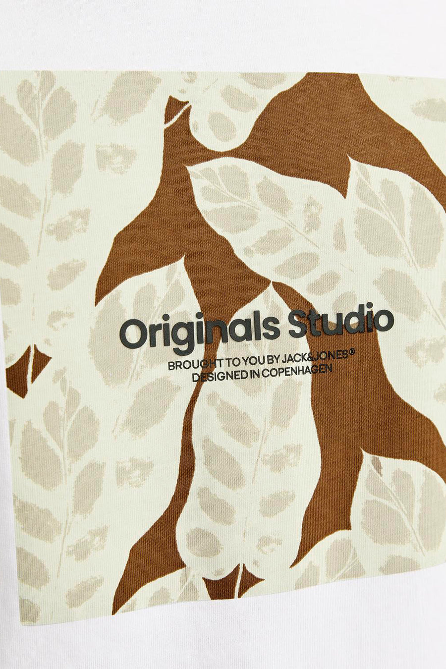 Detail van een wit shirt met bruin en crèmekleurig bladerpatroon en zwarte tekst 'Originals Studio'.