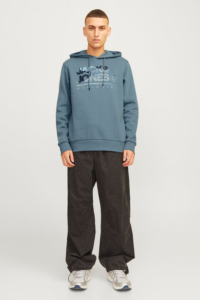 Man met blauwe hoodie, zwarte broek en zilveren sneakers staand op een witte achtergrond.