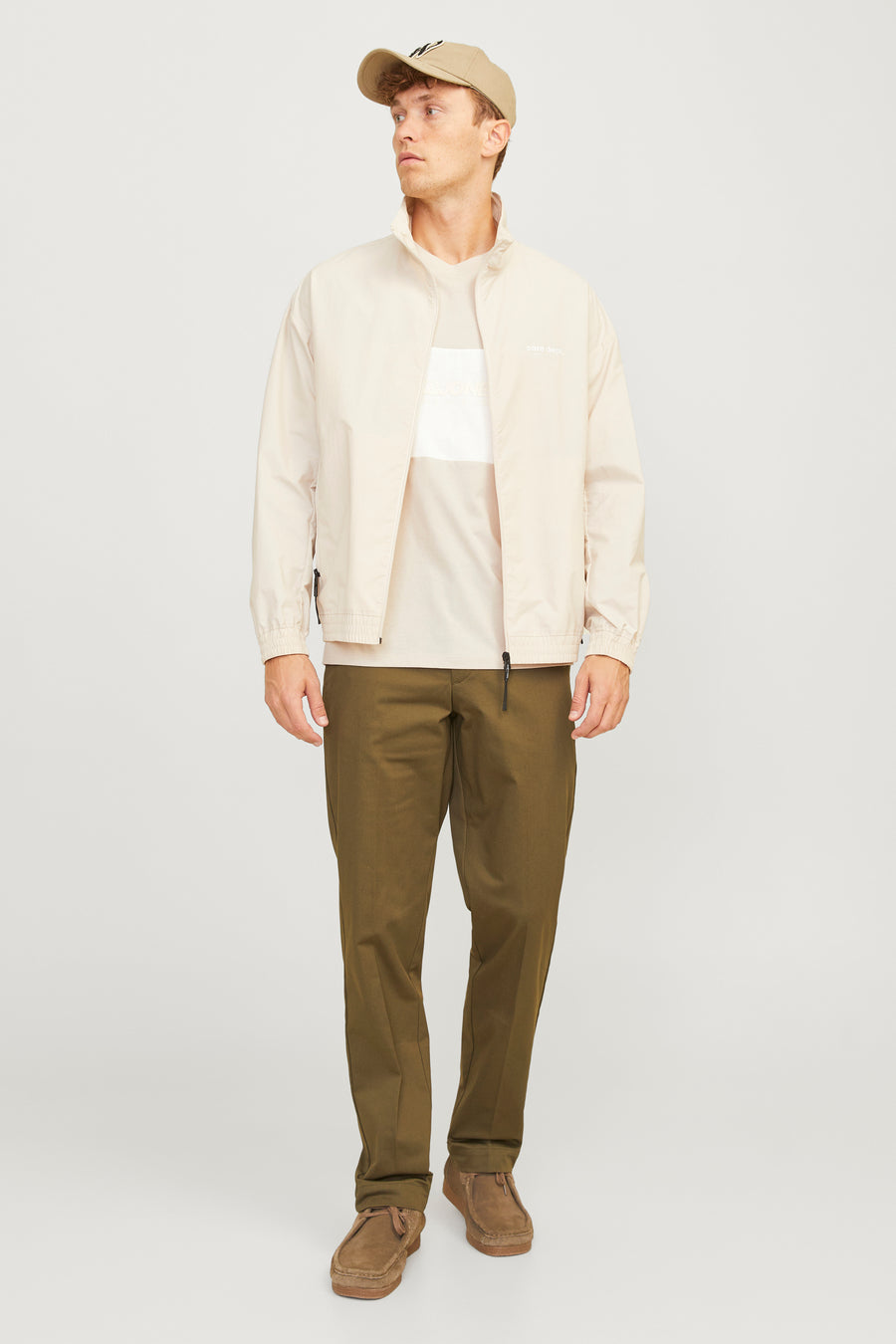 Man in beige jas, T-shirt, groene broek en pet, met bruine schoenen, staand tegen witte achtergrond.