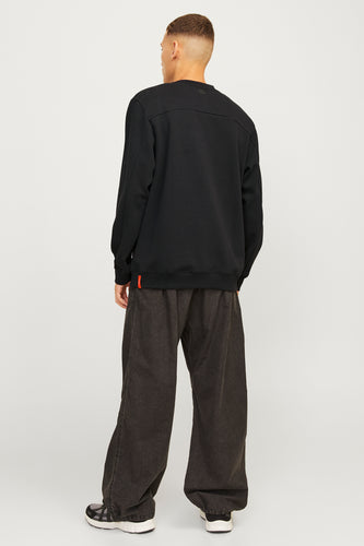 Man van achteren in zwarte trui en ruime, bruine broek tegen een witte achtergrond.