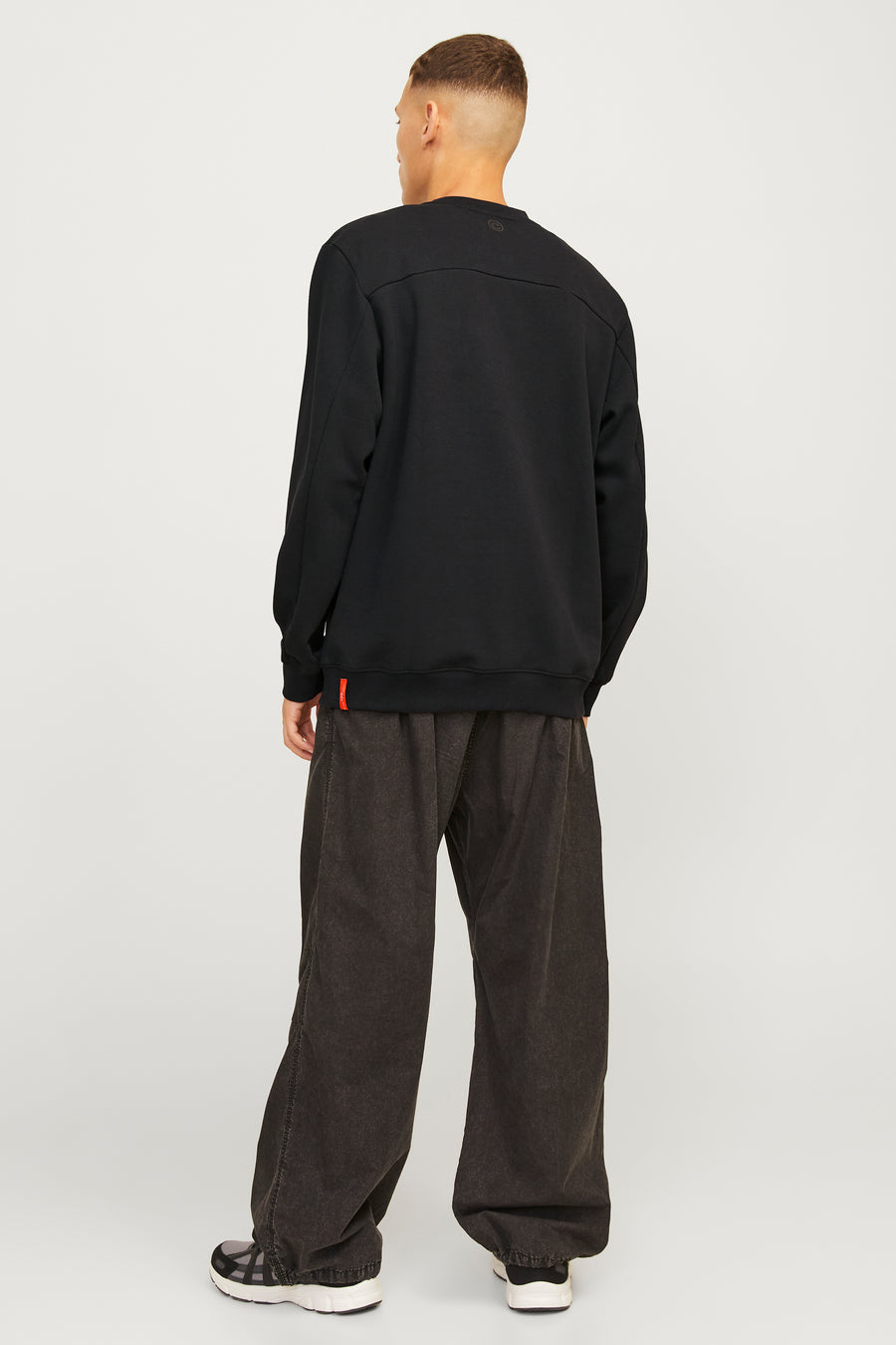 Man van achteren in zwarte trui en ruime, bruine broek tegen een witte achtergrond.