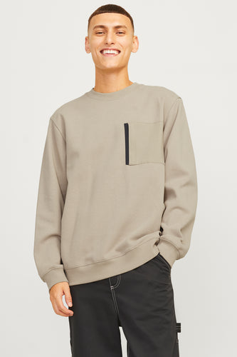 Man in taupe sweater met zak en zwarte broek met stiksels.