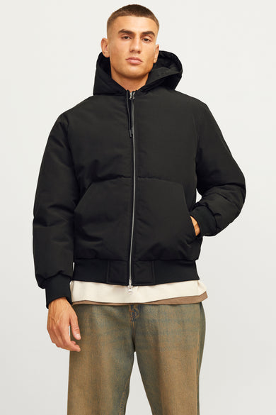 Manteau court noir de CORE BY JACK & JONES, avec capuche et porté sur un t-shirt beige et des jeans vert-brun.